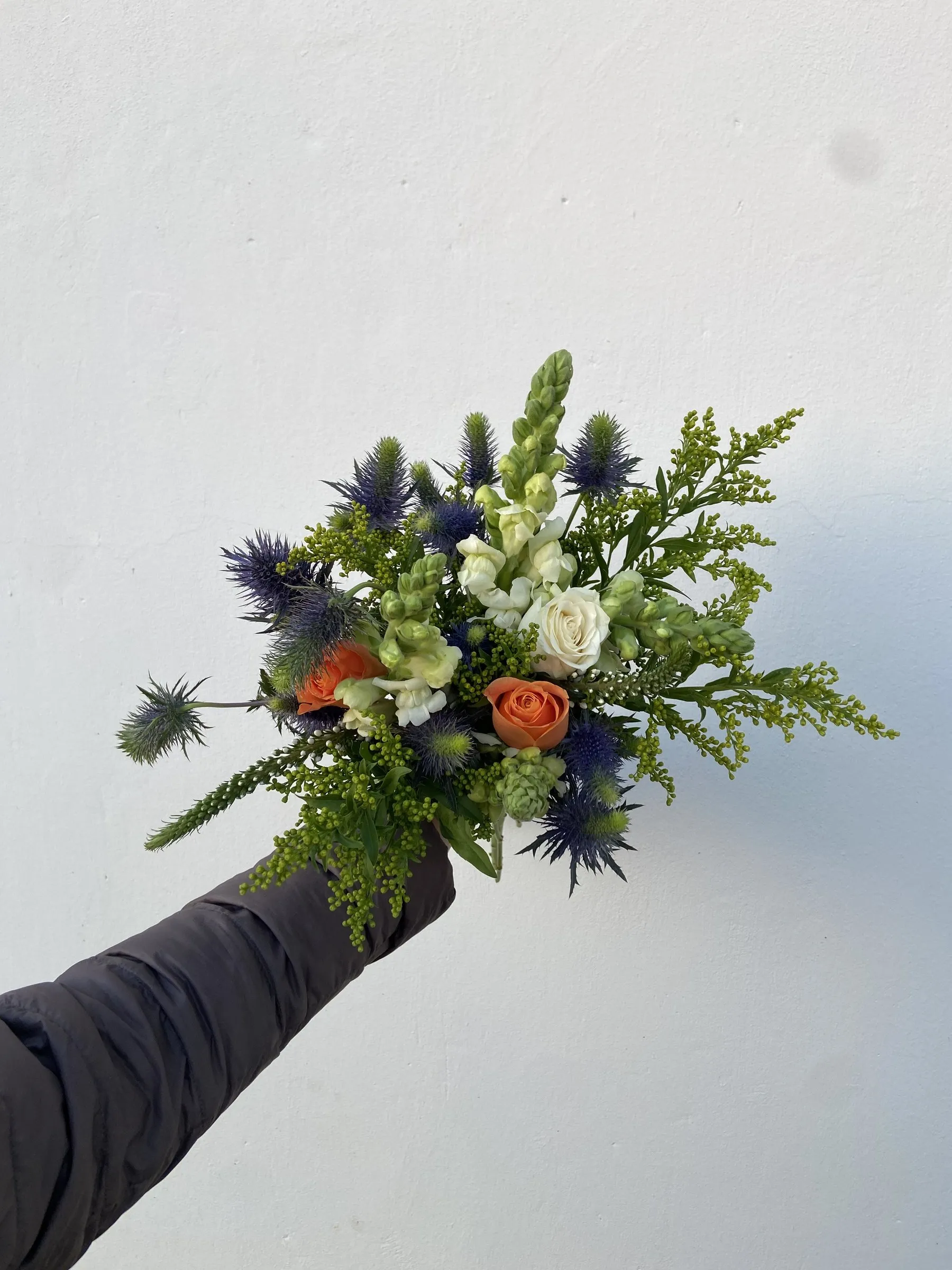 A hand holding out a bouquet of blue eryngium, white roses and foliage against the sky