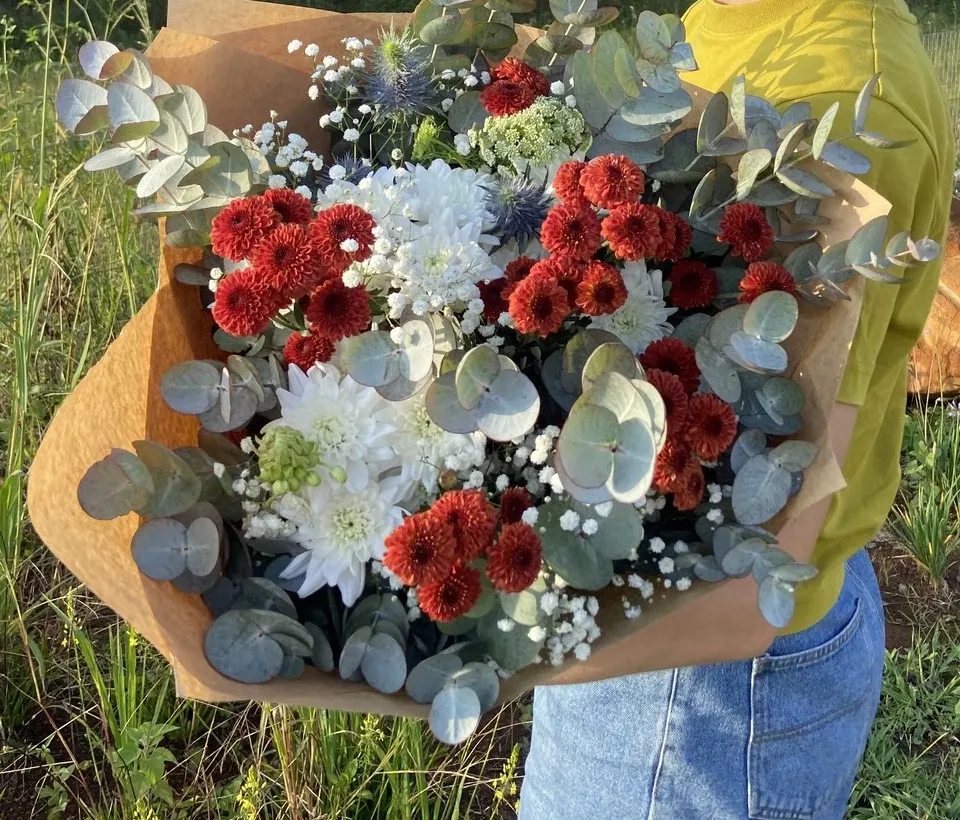 A large kraft-wrapped bouquet of red and white chrysanthemums, eryngium and eucalyptus, held in a field at golden hour