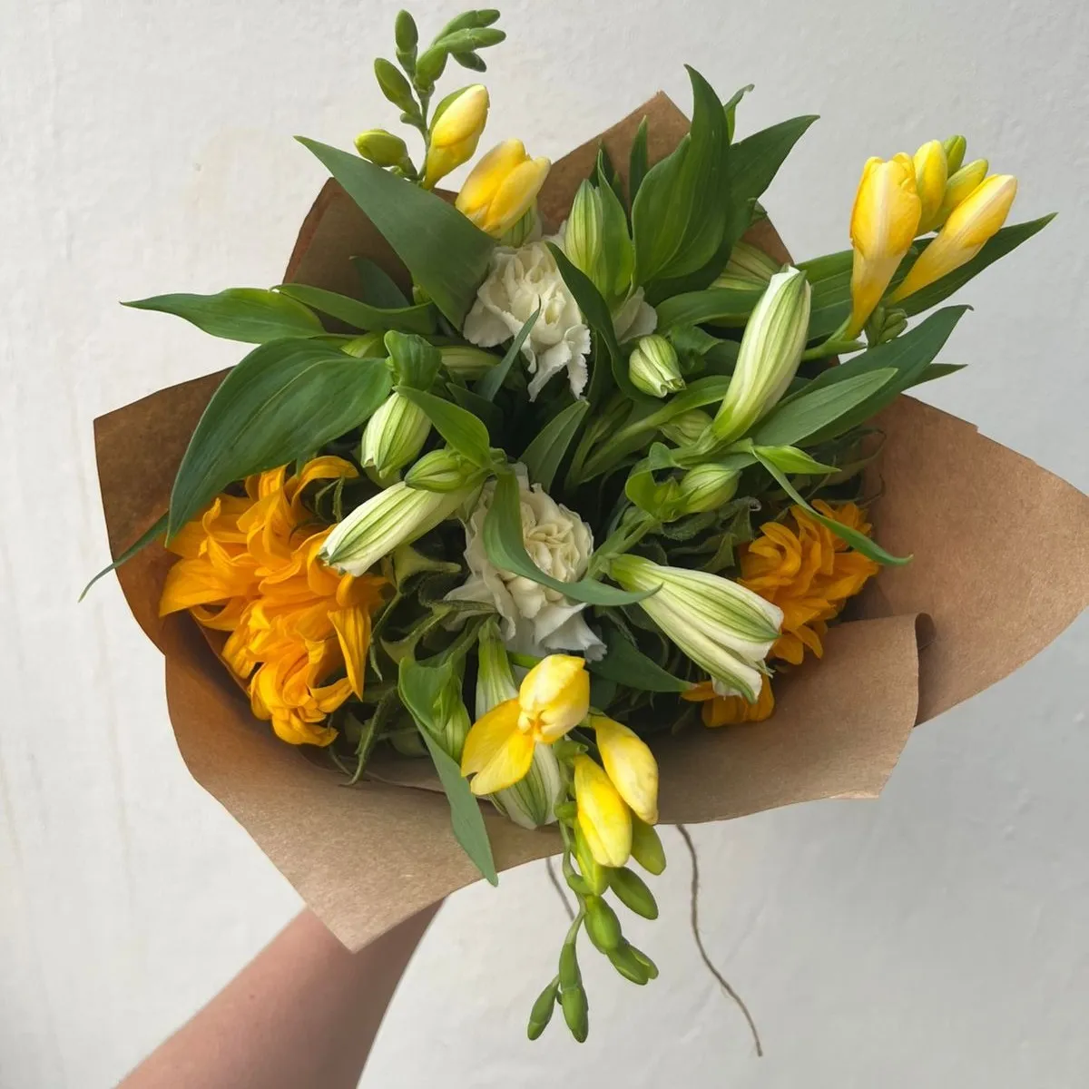 A kraft-wrapped bouquet of yellow freesias, cream carnations and orange chrysanthemums held against a white wall