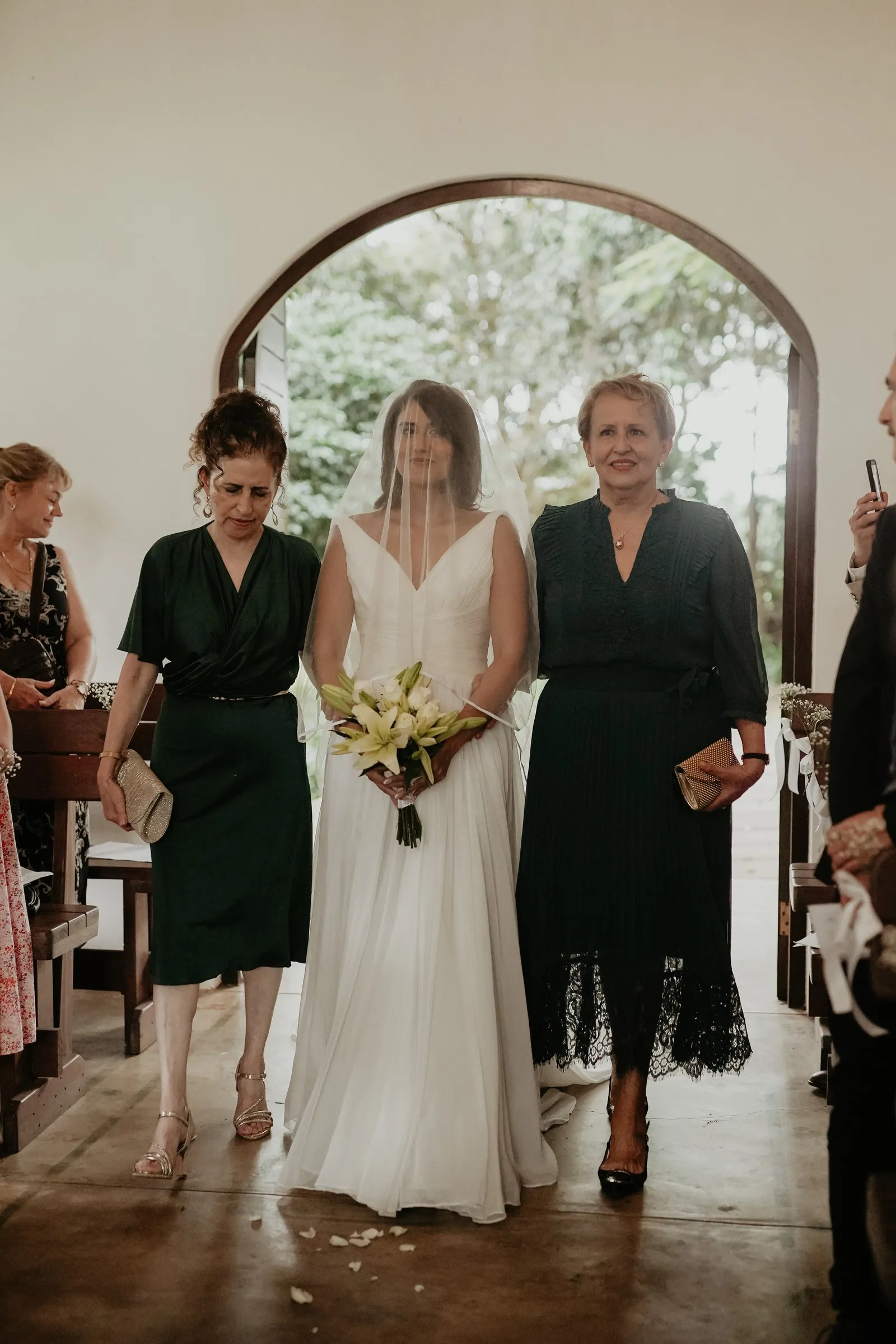 A bride walking down the aisle between two women, carrying a small white bouquet