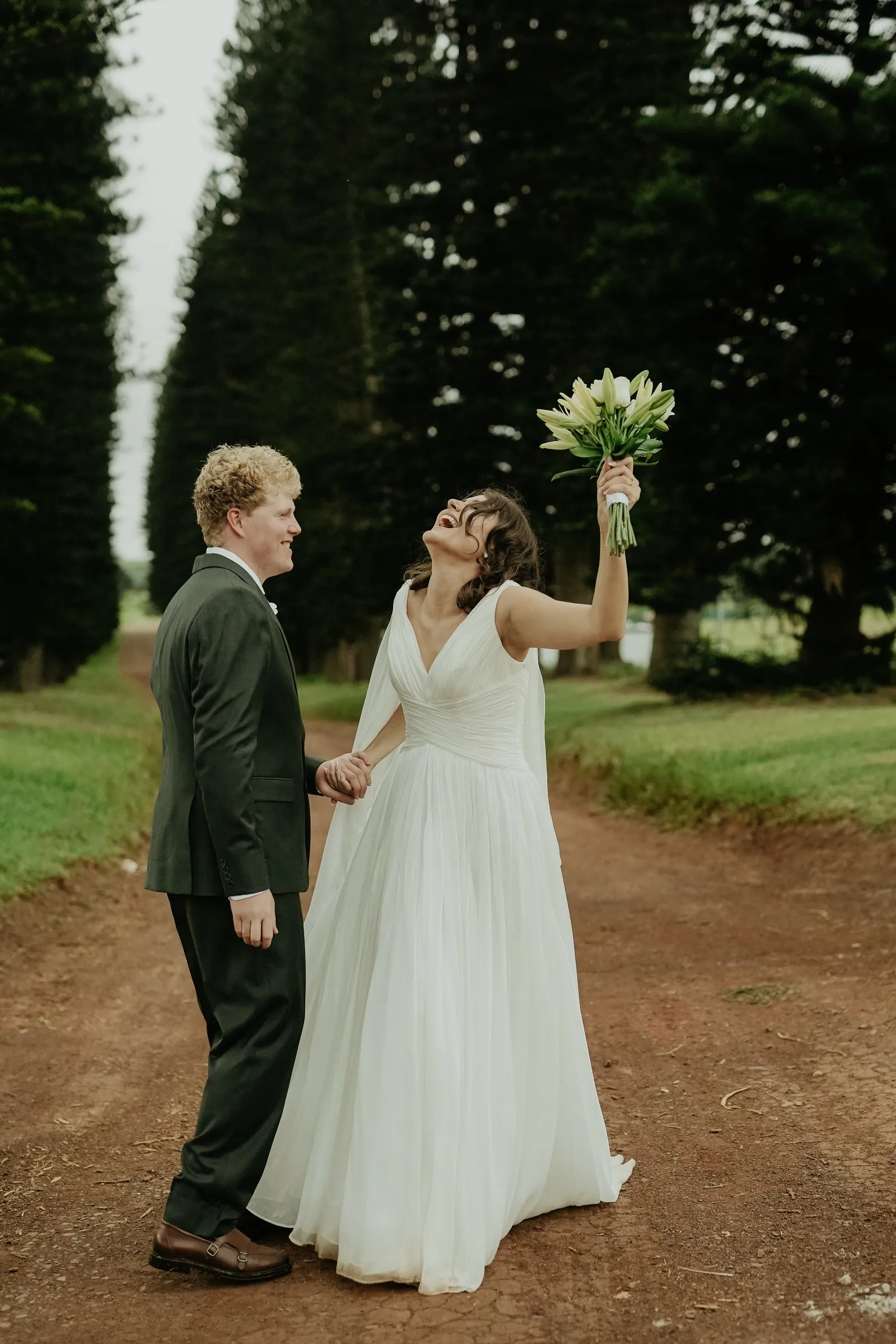 A bride raising her bouquet in the air on a tree-lined path, the groom beside her