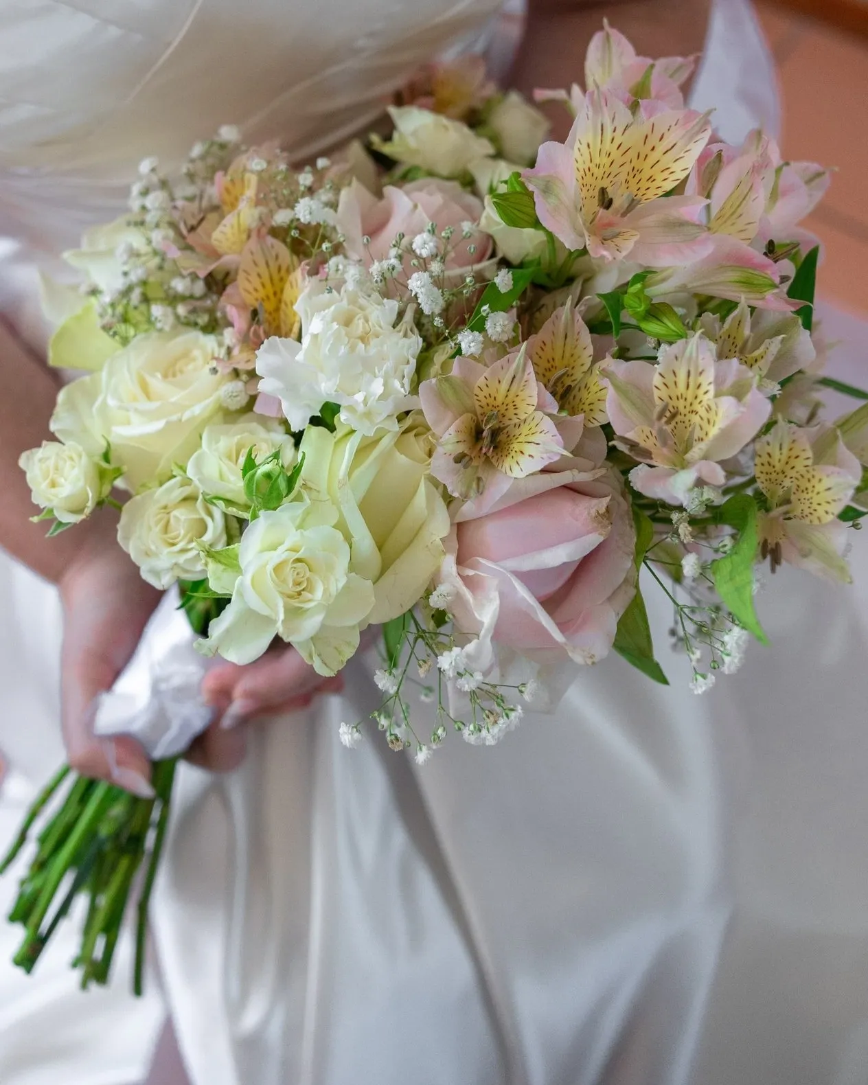 A cream and white bridal bouquet of roses and alstroemeria held against a satin gown