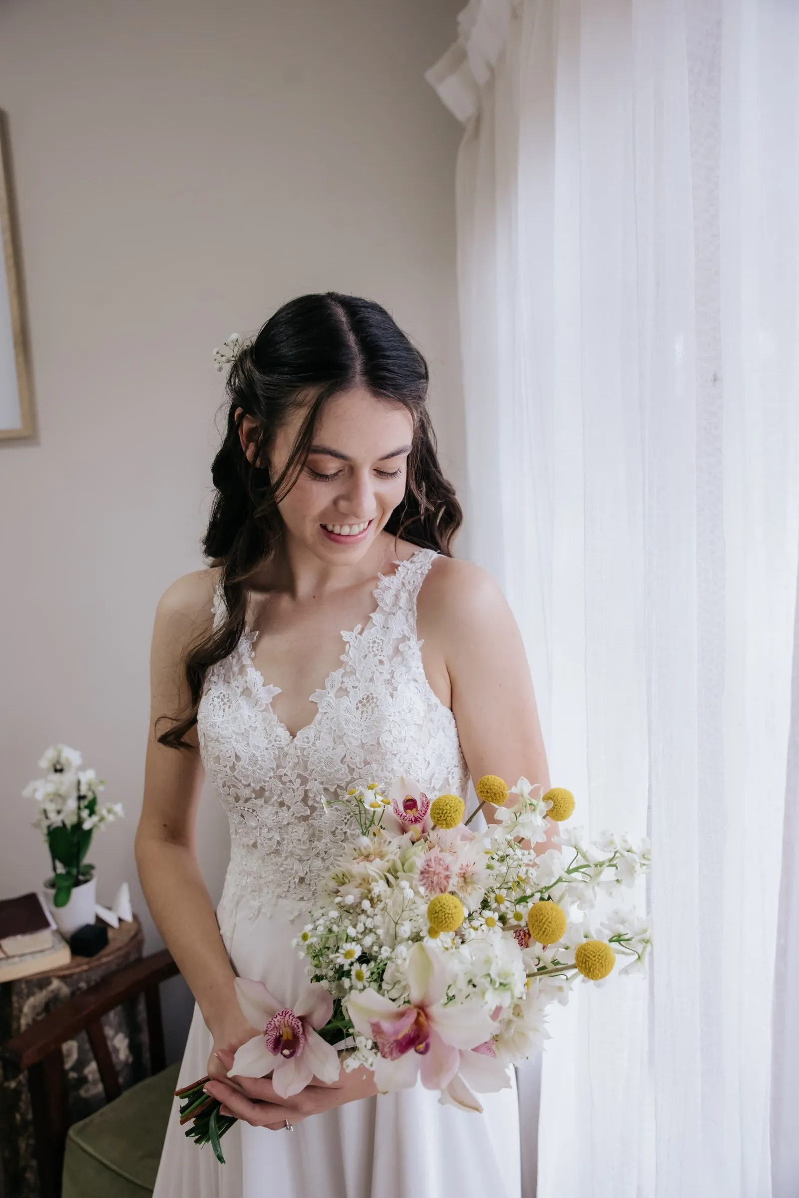 A bride in lace looking down at a bouquet of craspedia and white blooms