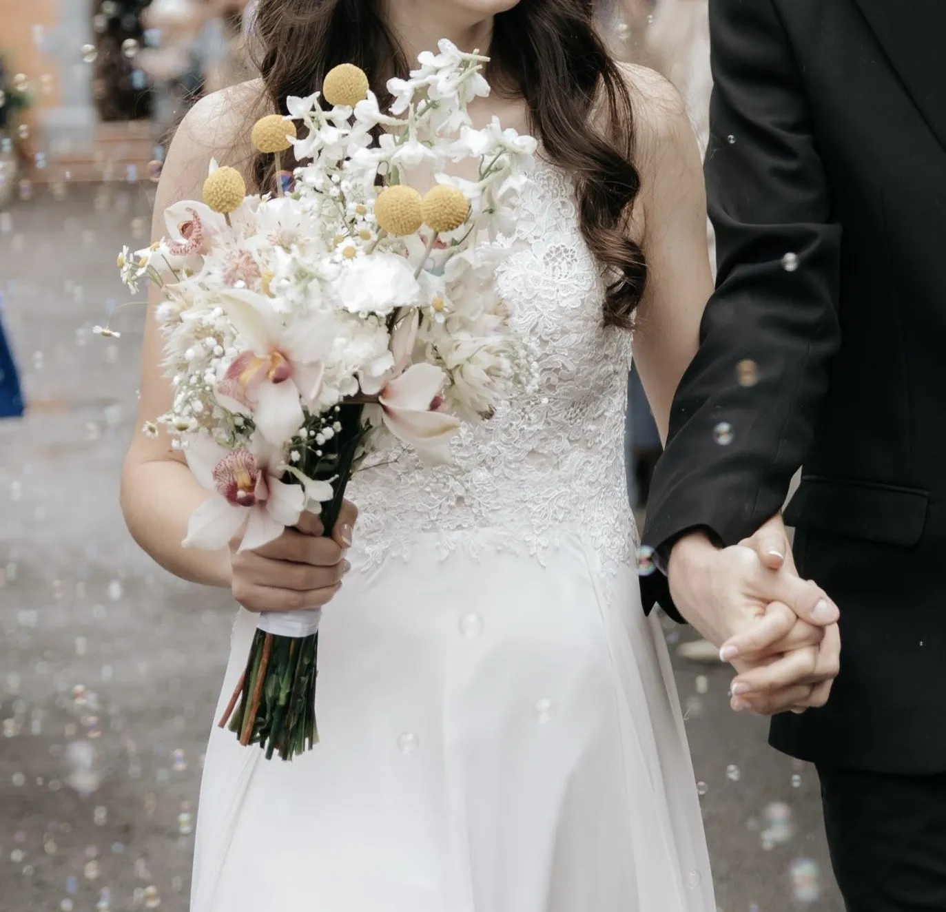 A couple holding hands through falling confetti, a soft pink and white bouquet between them
