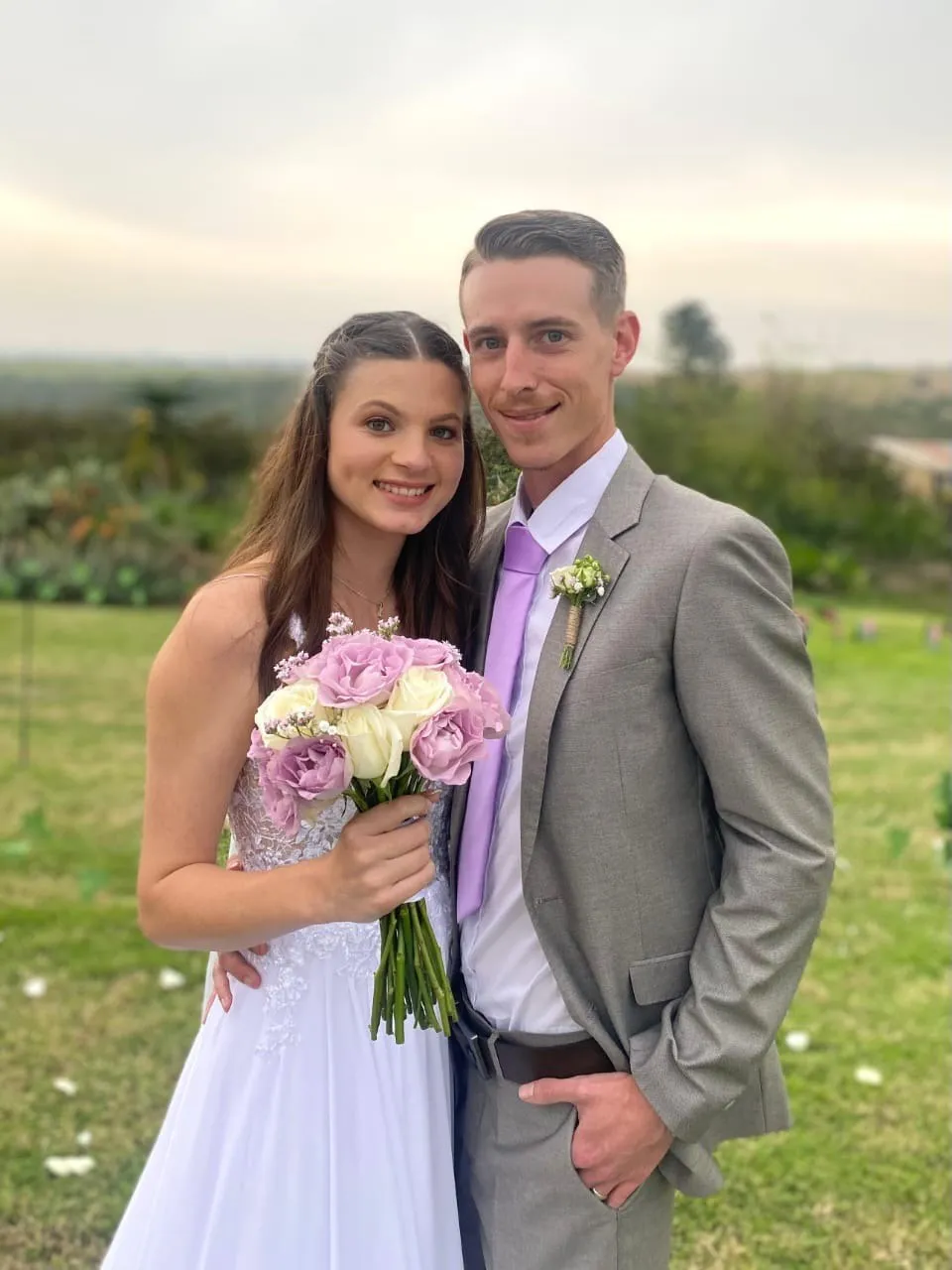 A couple standing together in a field, the bride holding a lilac and white bouquet