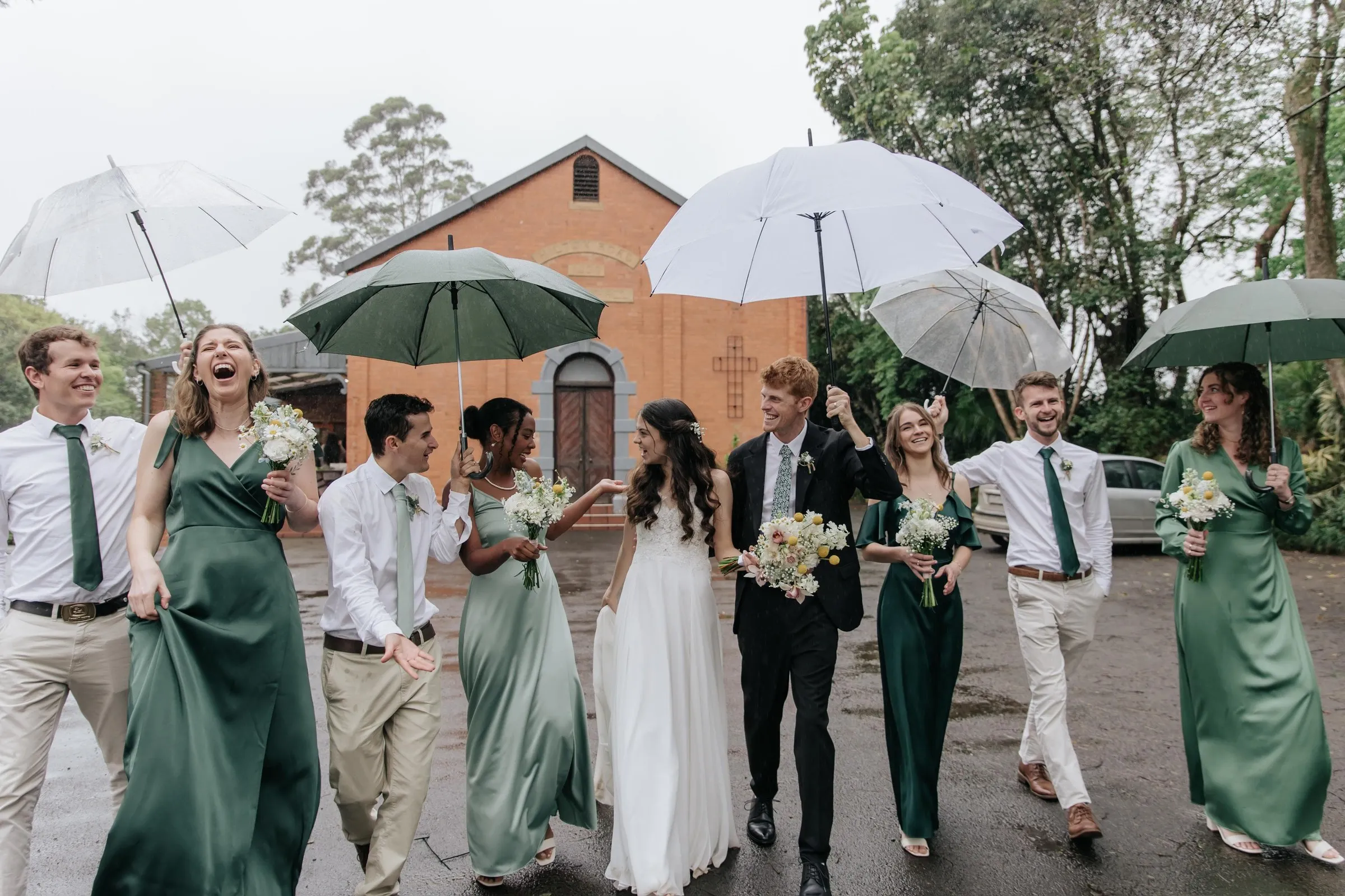 A wedding party walking under white umbrellas in the rain, bouquets held low
