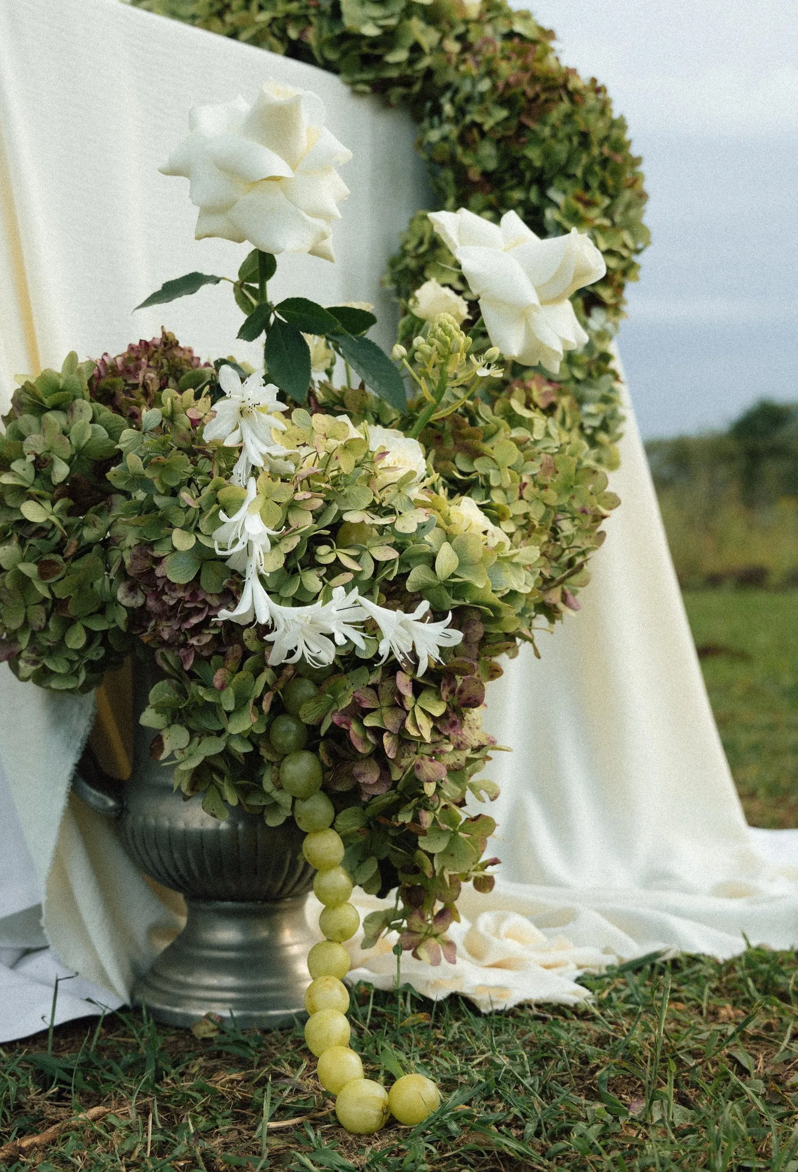 A green hydrangea and white lily arrangement spilling from a stone urn beside draped fabric